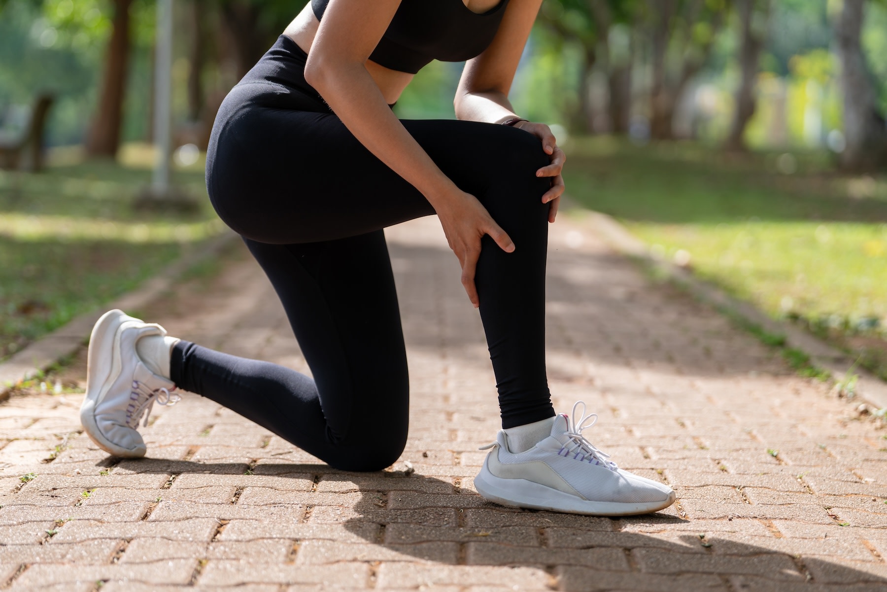 Close-up photo of a runner bent down on one knee on an outdoor path, scratching their calf and holding their knee.