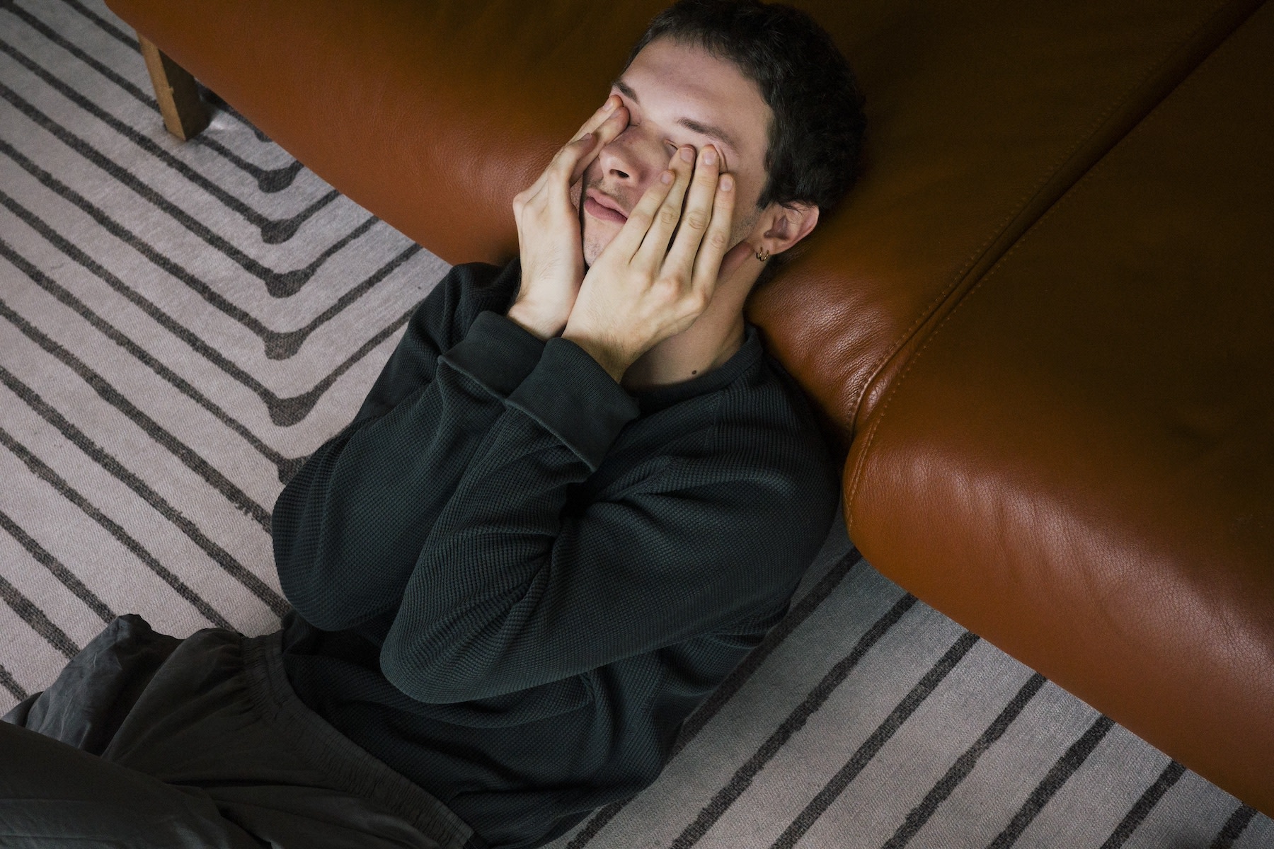 A man leaning against a couch at home while rubbing his eyes as he deals with the Sunday scaries.