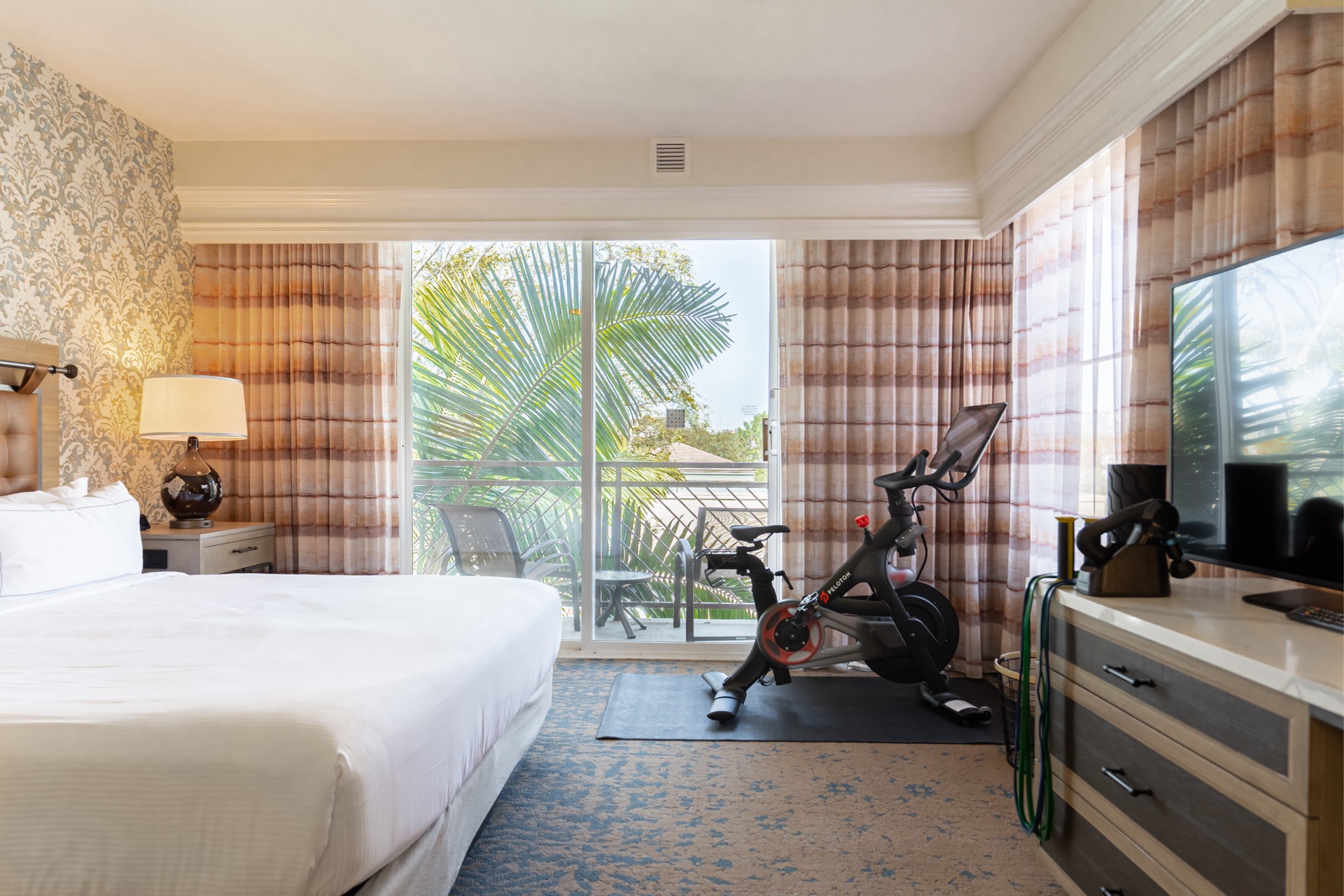 A Hilton hotel room with a bed, Peloton Bike, and recovery tools like a massage gun and roller on the TV stand