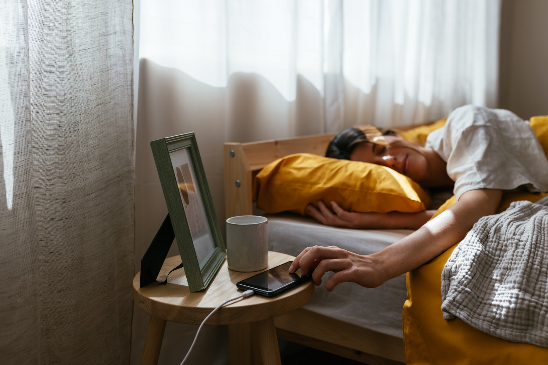 In this image for an article about how to fix your sleep schedule, a sleepy woman is lying in bed and reaching out to turn off the morning alarm on her phone. 