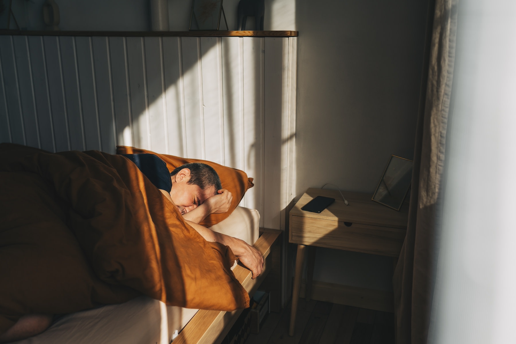 A man who is waking up tired in bed after a full night of sleep. He's lying in a modern-looking bed looking disgruntled.