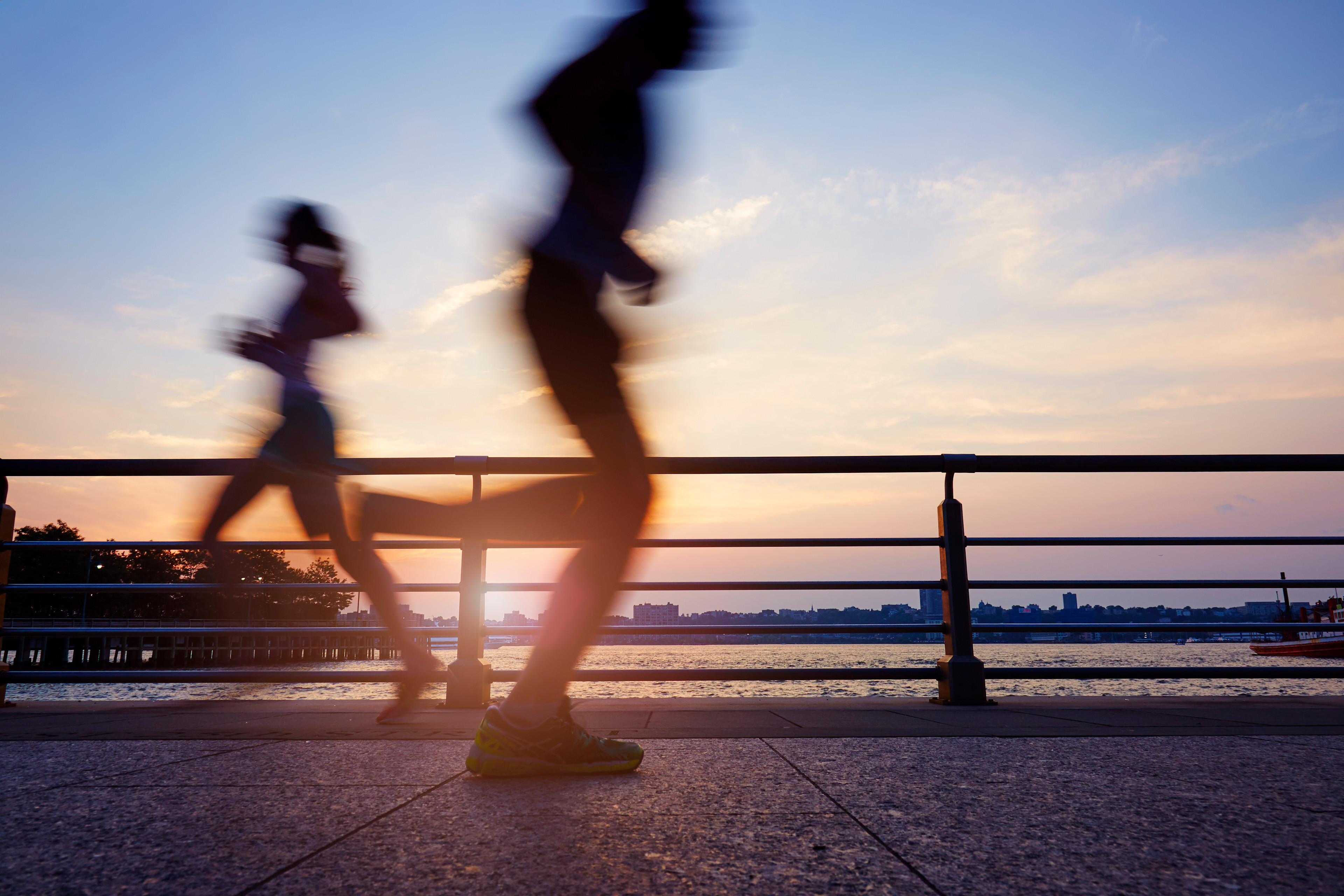The silhouettes of two runners on a waterside path during sunrise to illustrate the concept of running twice a day.