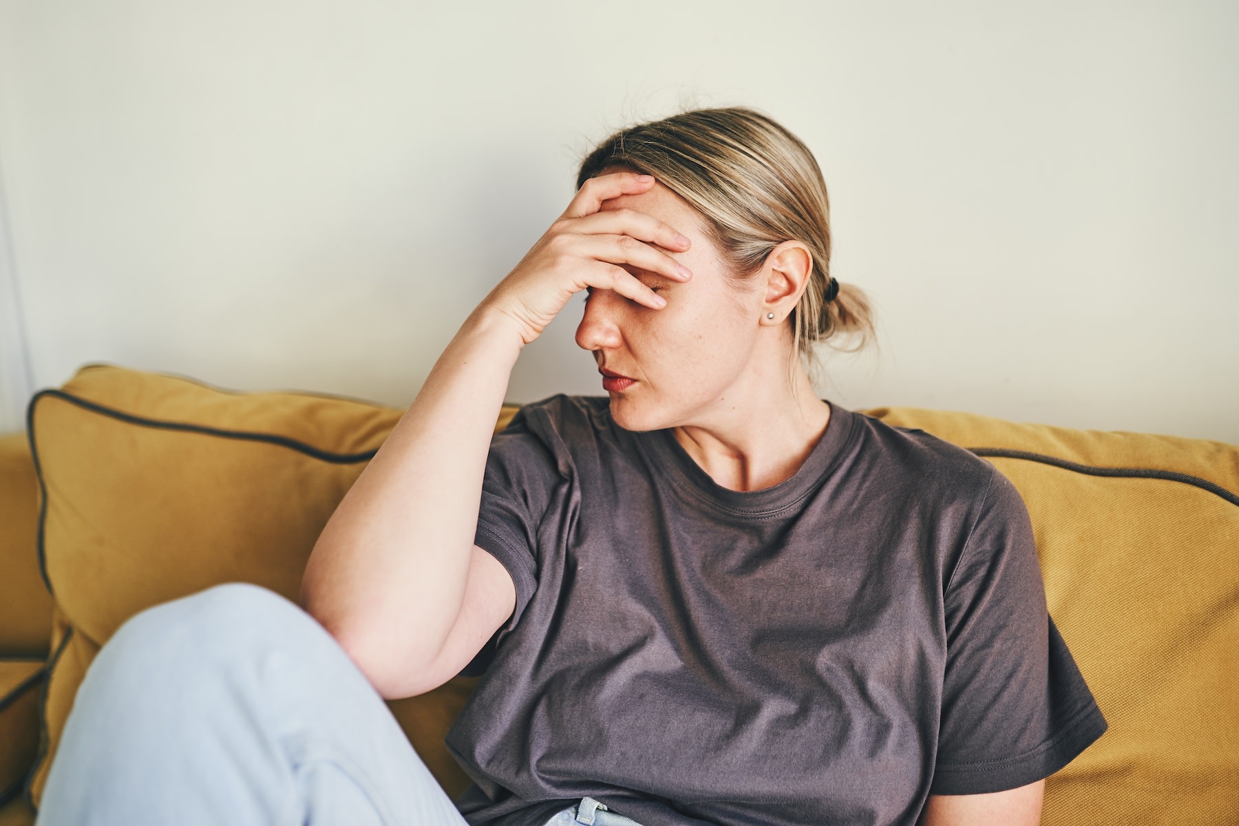 A mentally exhausted woman sitting on the couch looking stressed. She is holding her head in her hand.