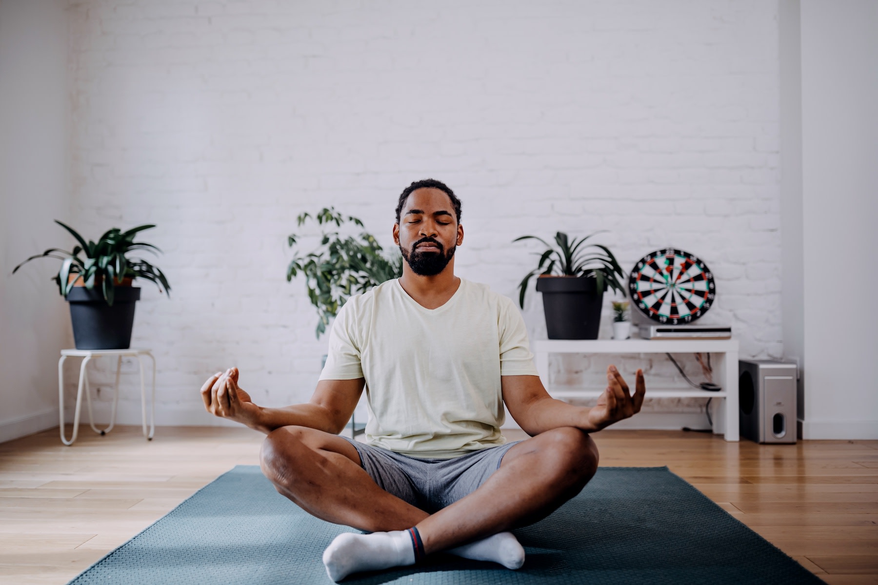 A man practicing mantra meditation. He's sitting on a mat in a sunny room with his eyes closed, sitting in a criss-cross position with his hands resting on his knees.