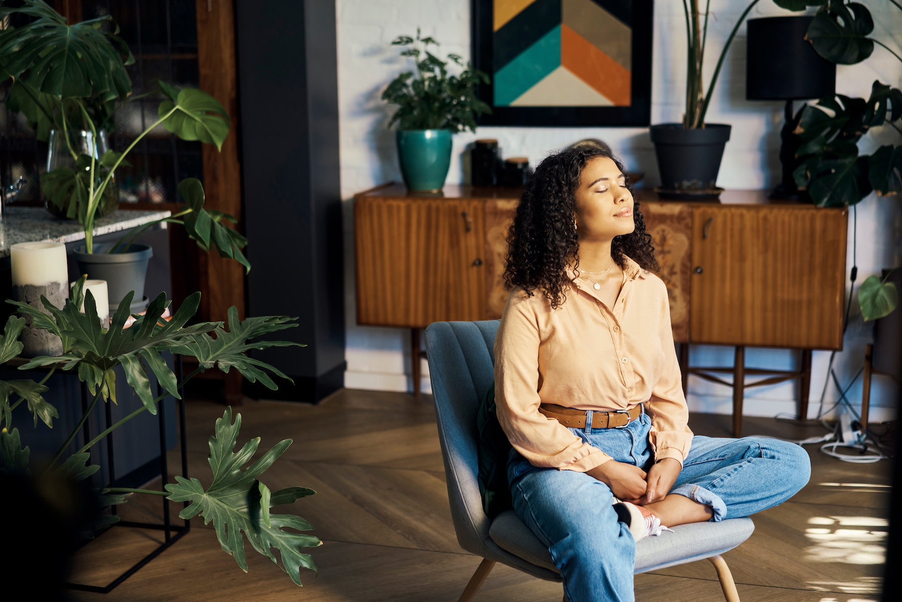 A woman sitting on a chair in a sunny room at home while practicing a body scan meditation.