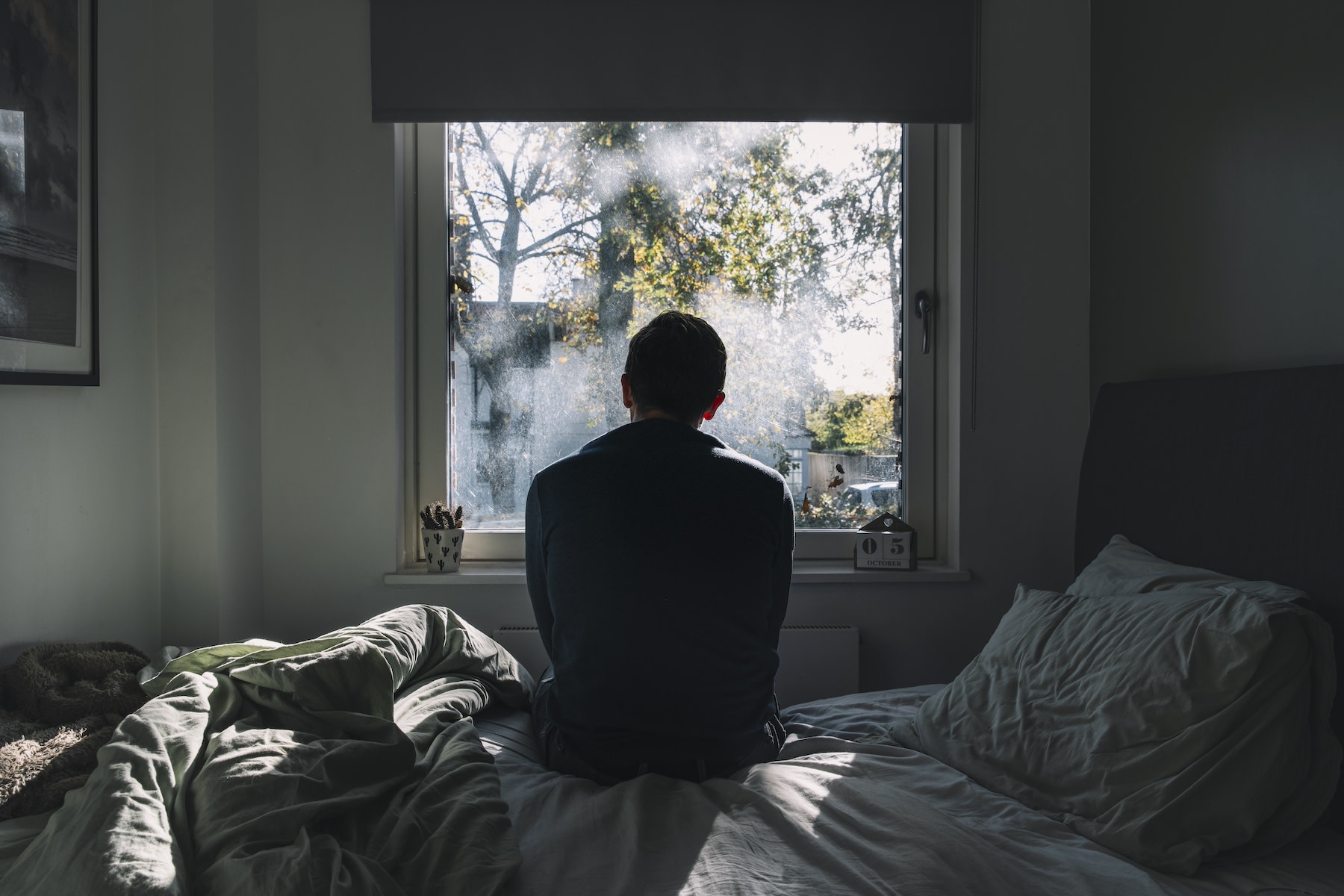 A rear-view photo of an unrecognizable man sitting up in bed and staring out the window after waking up with anxiety.