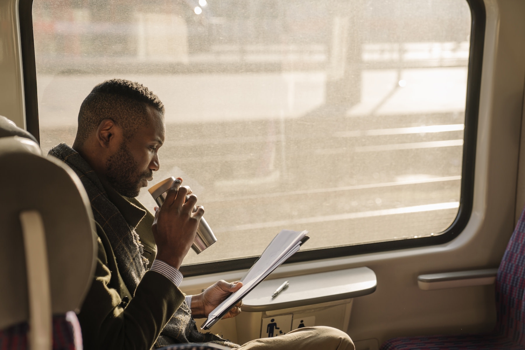 A businessman sipping coffee from a thermos while reading documents while sitting on a sunny train.