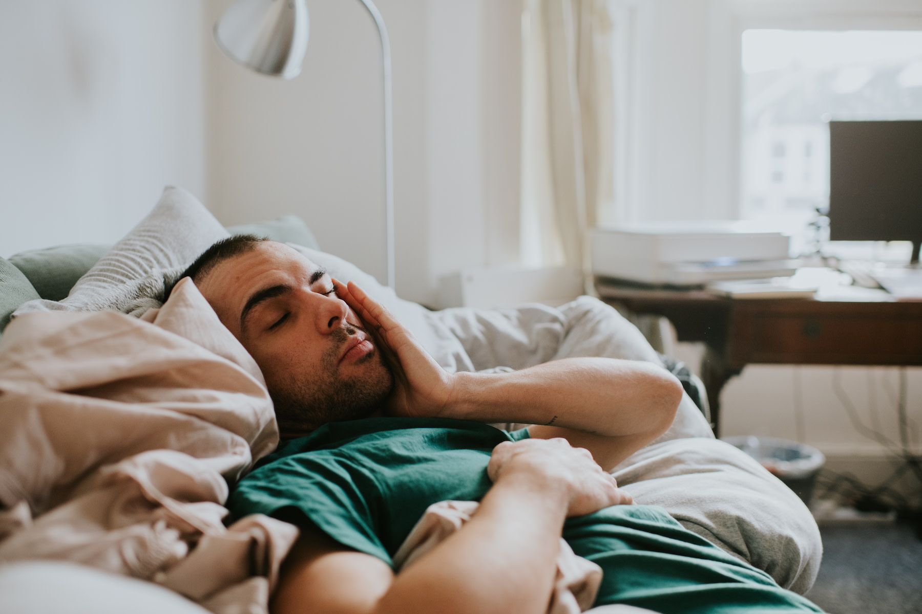 A tired man lying in bed while rubbing his face as he suffers from social jet lag.