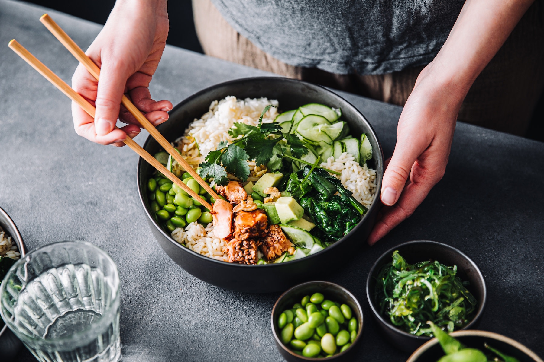 A person eating a poke bowl with chopsticks. The bowl has protein sources like salmon and edamame.