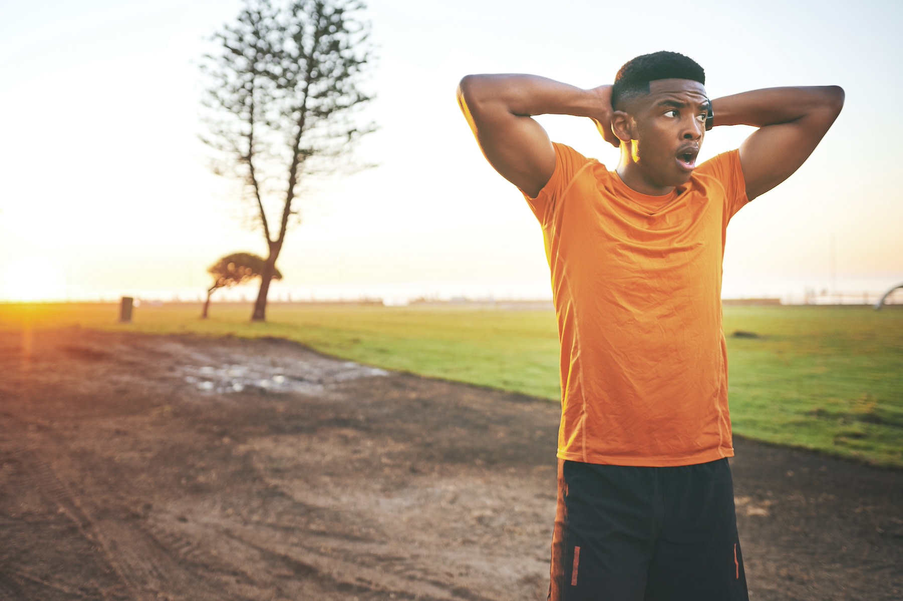 A man yawning during a workout. He is standing with his arms on his head and his mouth open outdoors.