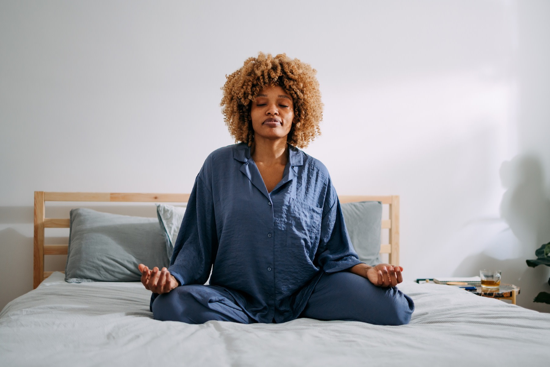 A woman practicing daily meditation while sitting on her bed in a sunny bedroom.