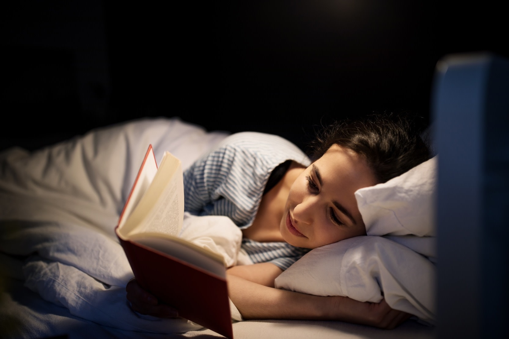 A woman reading a book while lying in bed at night as part of her bedtime routine.