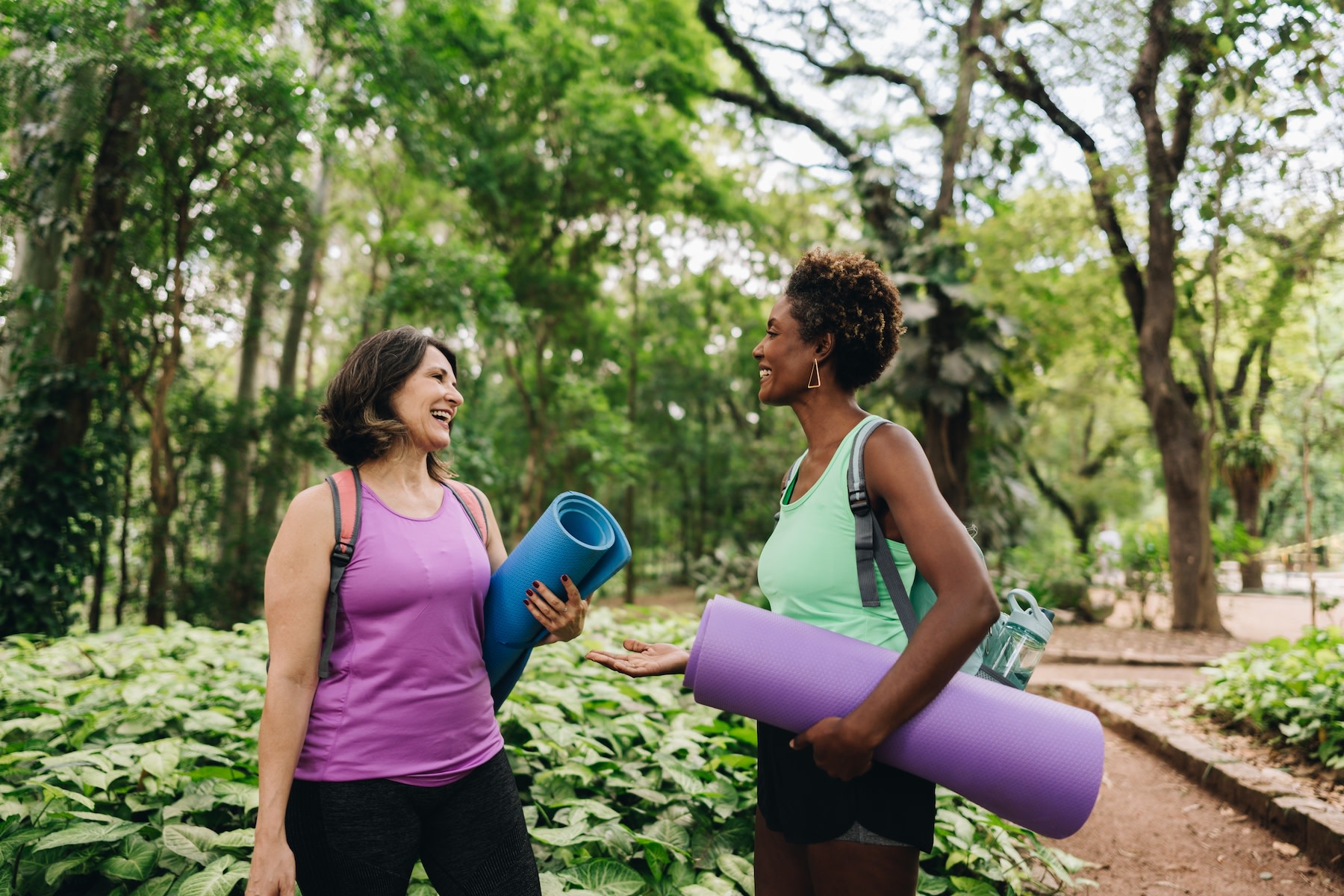 Two women happily talking outside while holding yoga mats. Exercising and spending time with friends are great self-care ideas.