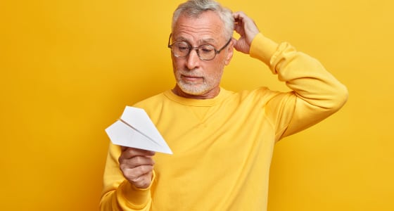 Man wearing glasses holds paper airplane while scratching head.