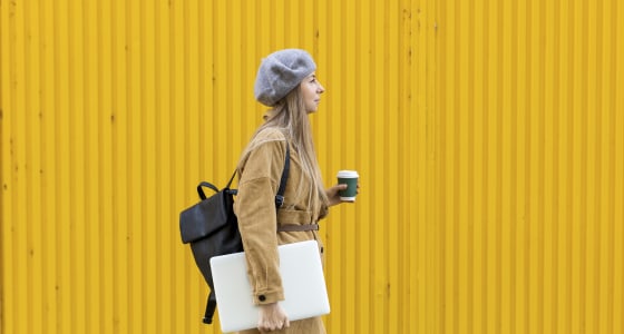 Woman holding coffee and laptop walking down the street.