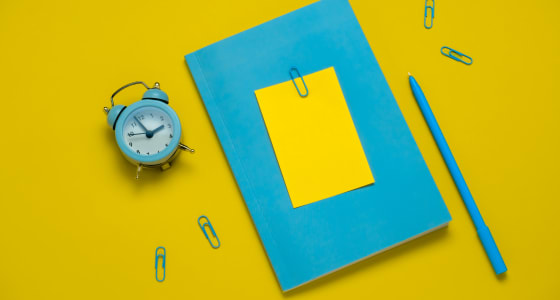 Blue notepad, pen and alarmclock on a plain yellow background