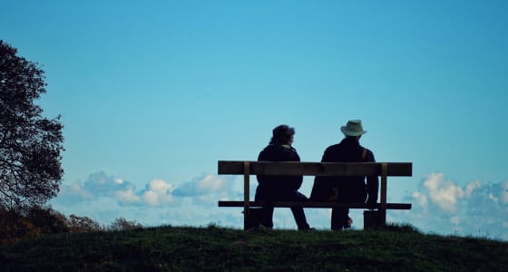 A couple sitting on a bench facing away from the viewer win a countryside setting with blue skies