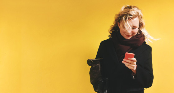 Woman looking at her phone, dressed in dark clothing standing in front of a yellow background.