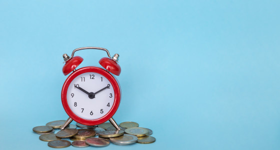 Red alarm clock sitting on a small pile of coins infront of blue background