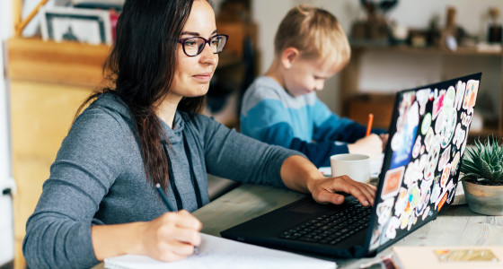 A woman working from home, using her laptop, with a young child drawing beside her.