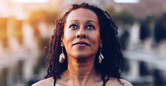 A middle-aged woman of African descent wearing her hair in twists with dangly earrings and purple lipstick.