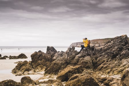 Woman sat on beach rocks, wearing yellow jacket, uses binoculars.