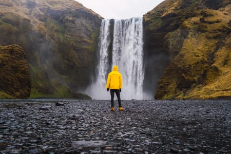 Man standing, wearing yellow jacket, in front of waterfall.