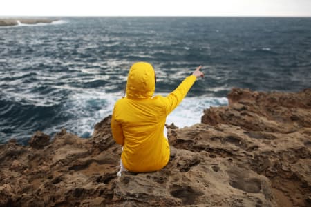 Woman sat on a rock, wearing yellow jacket, in front of the sea.