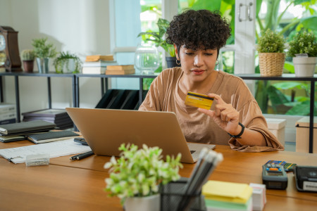 Woman holding yellow card, in front of computer, surrounded by green plants.