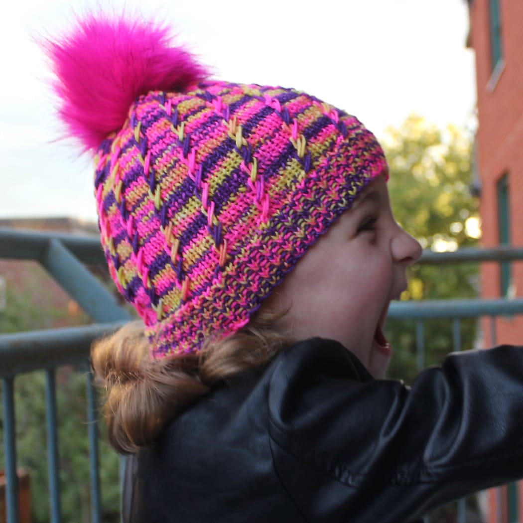 Side view of child wearing bright yellow, pink, and purple toque with pink pompom and spiralling criss-crossing surface texture.