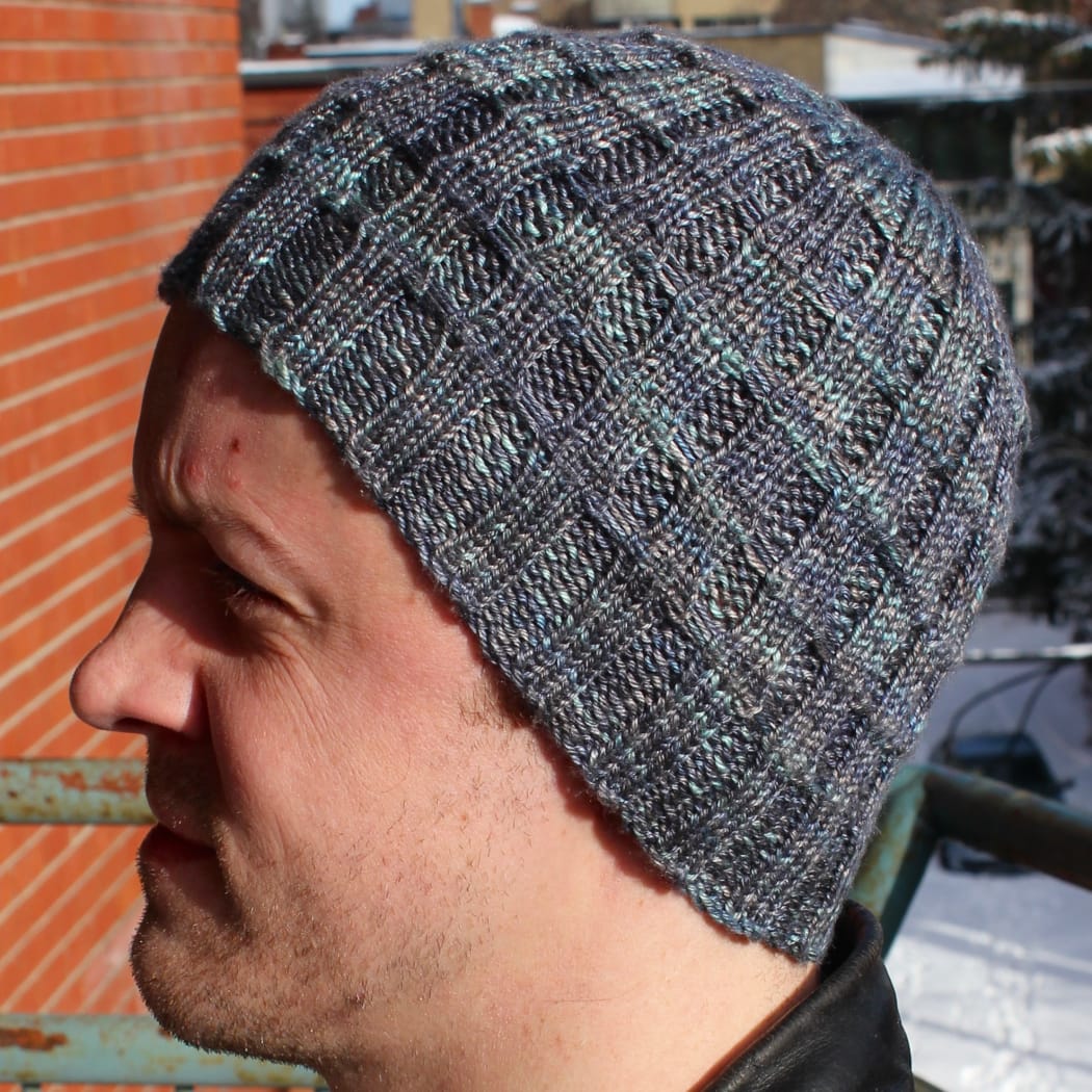 Man wearing grey-blue hat with ribbing crossed by parallel lines.