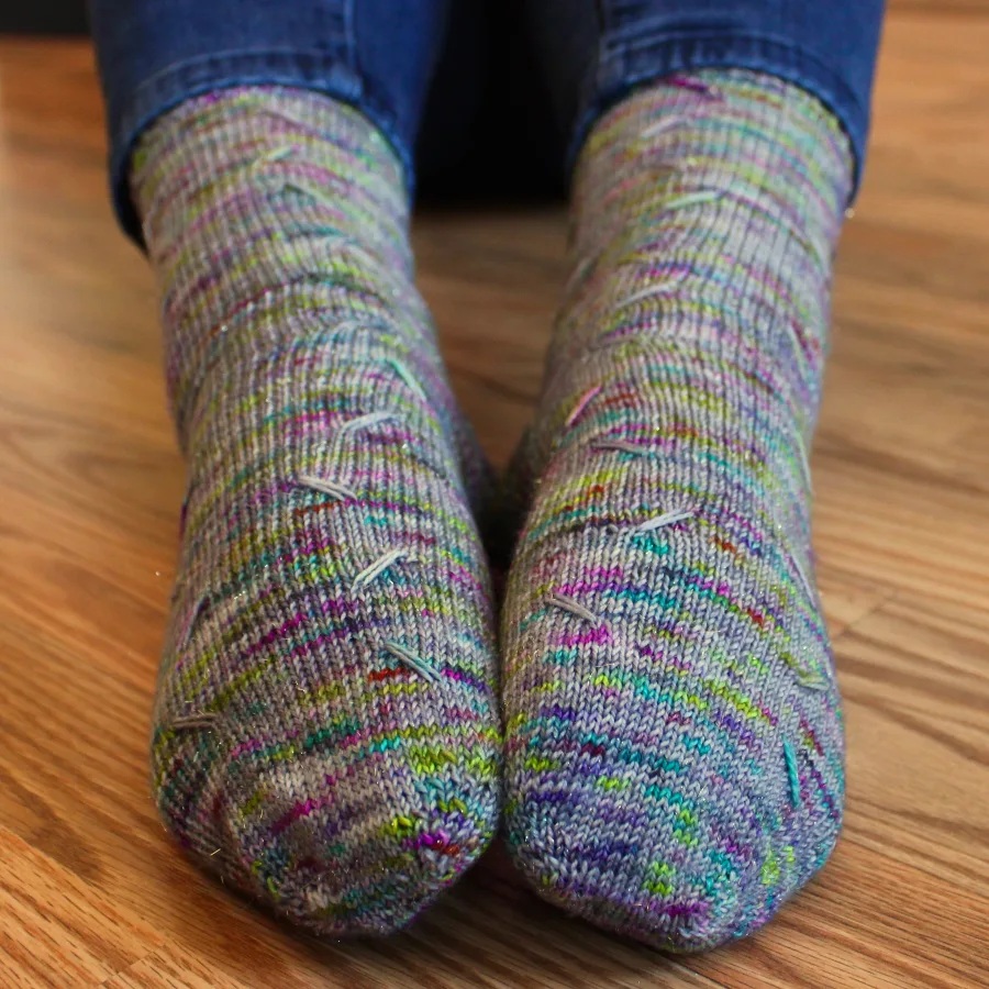 Toe up view of feet wearing grey socks with rainbow speckles that have lines of diagonal elongated stitches in two columns.