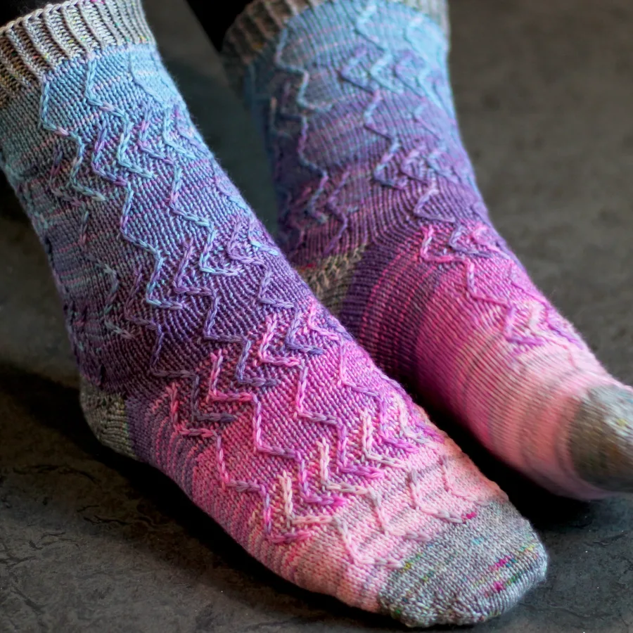 Side view of feet wearing pink, purple, and blue knitted socks with grey heels, toes, and cuffs; they have an overlapping zigzag surface texture.