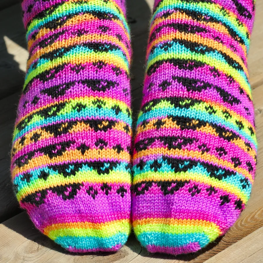 Close up of toes of stranded colourwork socks with black 8-bit sunglasses and sparkles motif on neon rainbow striped background.