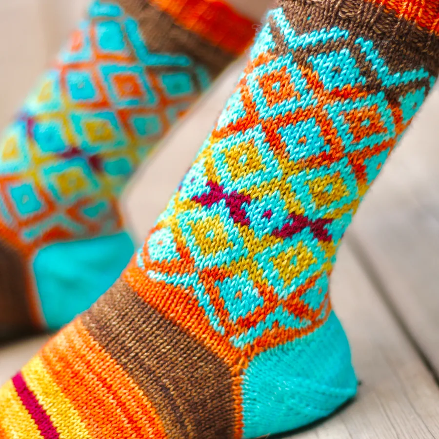 Close up side view of feet wearing brown, yellow, orange, and red striped knitted sock with overlaid bright blue diamonds motif on leg and bright blue heel.