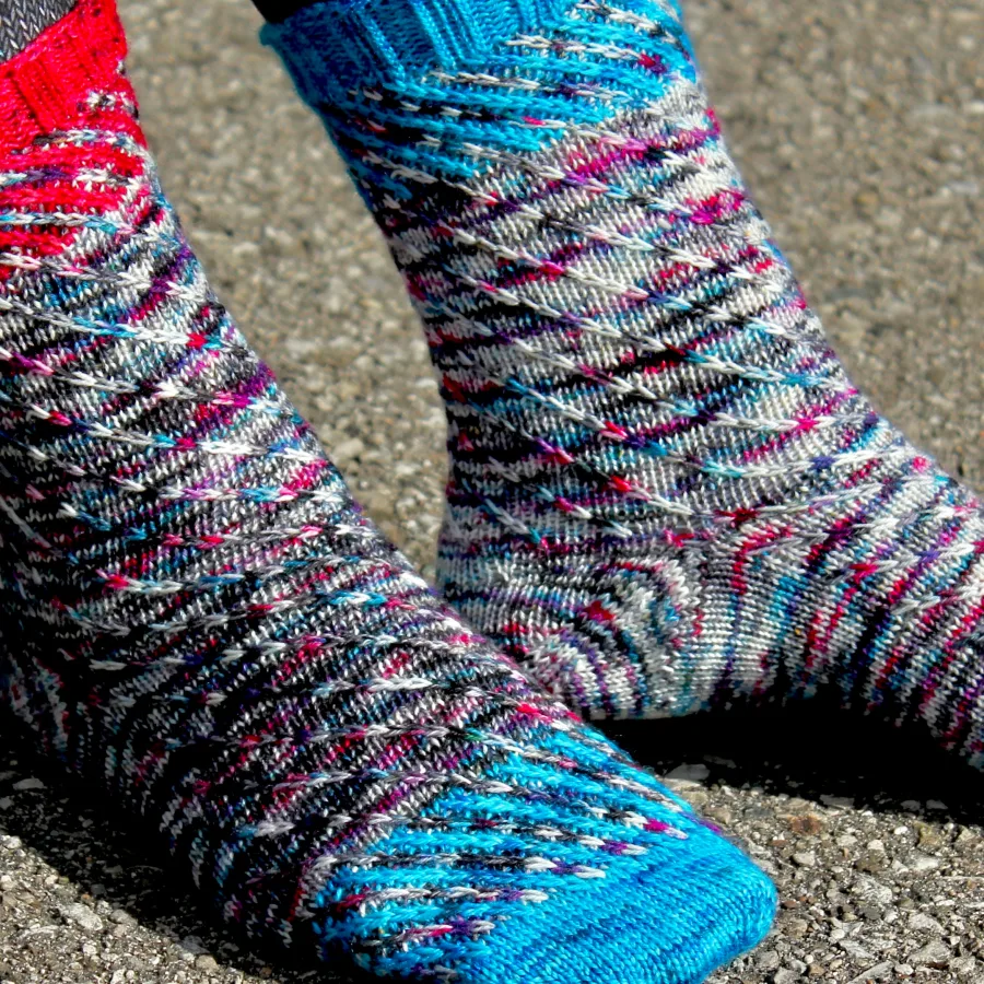 Side view of feet wearing knitted socks in a wildly-speckled yarn with a spiralling slipped-stitch texture; the socks have diagonal accent-colour sections at the toes and cuffs.