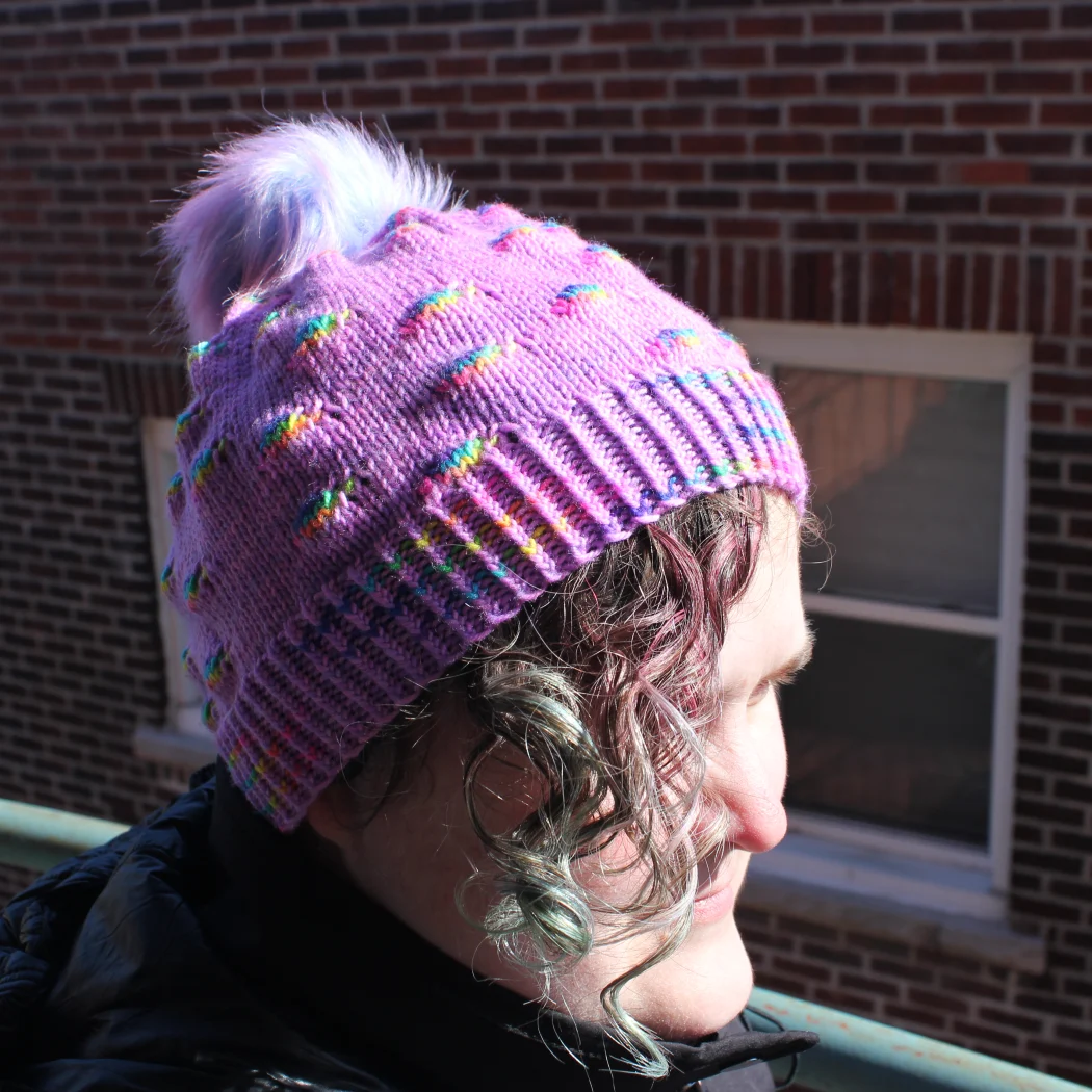 Woman wearing bright purple hat with pompom and randomly-distributed rainbow sections.