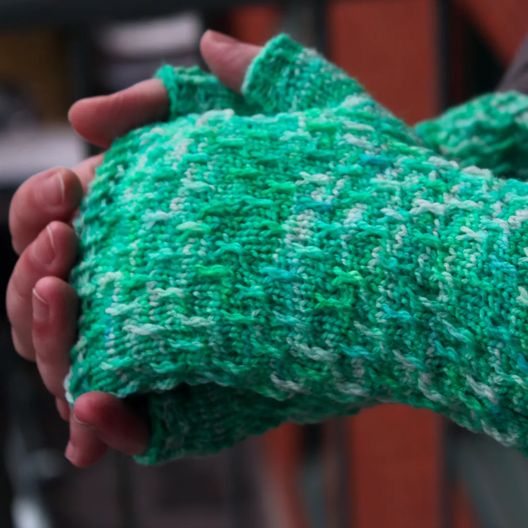 Side view of hands held together wearing bright green, white, and blue fingerless mitts with twisted surface detail.