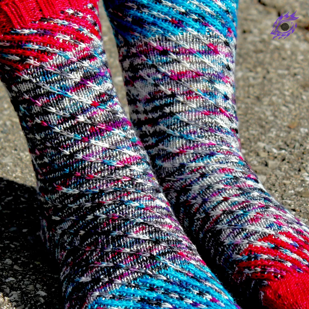 Feet wearing knitted socks in a wildly-speckled yarn with a spiralling slipped-stitch texture; the socks have diagonal accent-colour sections at the toes and cuffs.