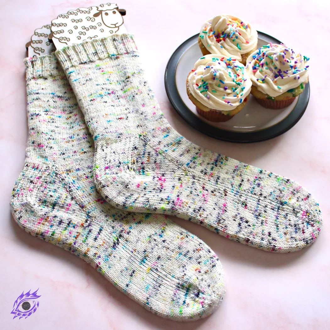 Wide view of rainbow speckled socks on sock blockers beside a plate of rainbow cupakes.