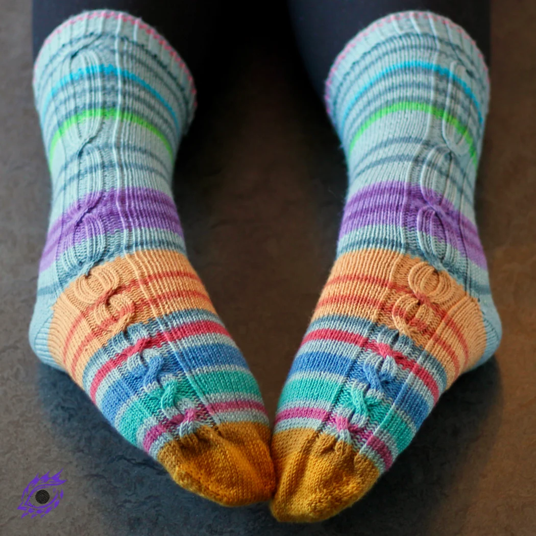 Feet wearing striped knitted socks that have gradually-lengthening cables in panels on the outsides of the socks.