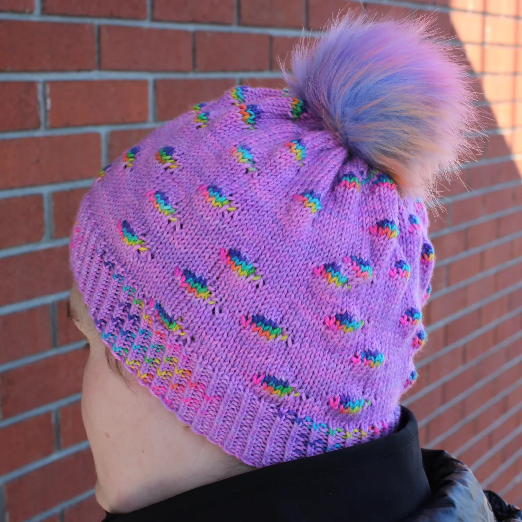 Woman wearing bright purple hat with pompom and randomly-distributed rainbow sections.