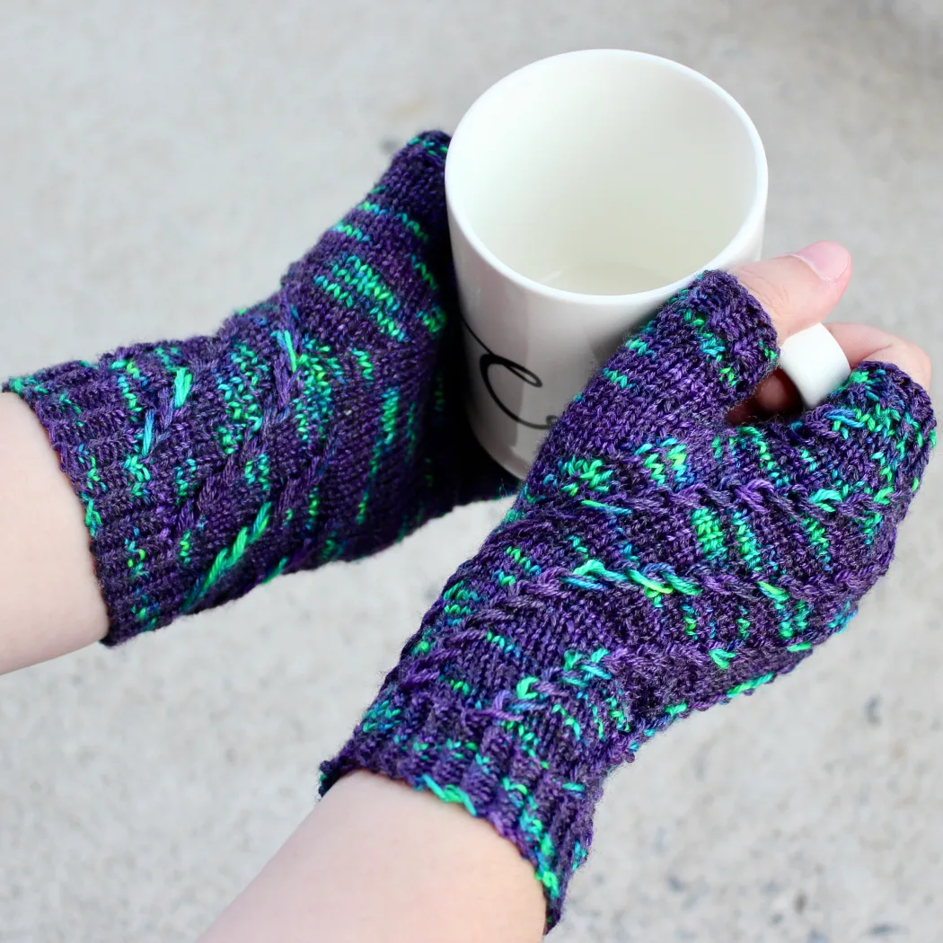 Top-down view of hands holding a white mug and wearing dark purple fingerless mitts with bright green flecks in a twisting spiralling pattern.