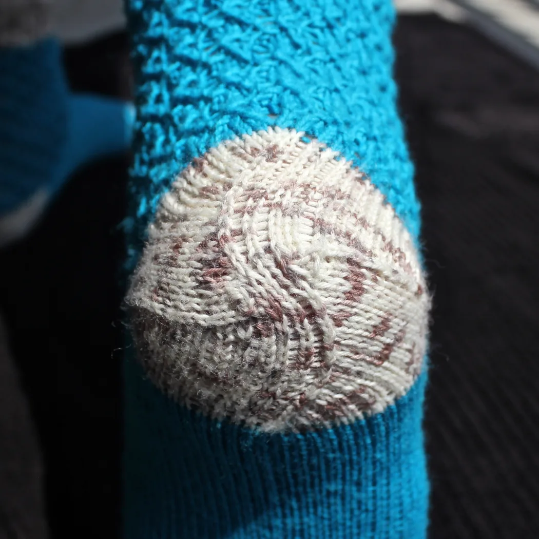 Close-up of reinforced spiral afterthought heel knit in white and brown speckled yarn on a bright blue lace sock.