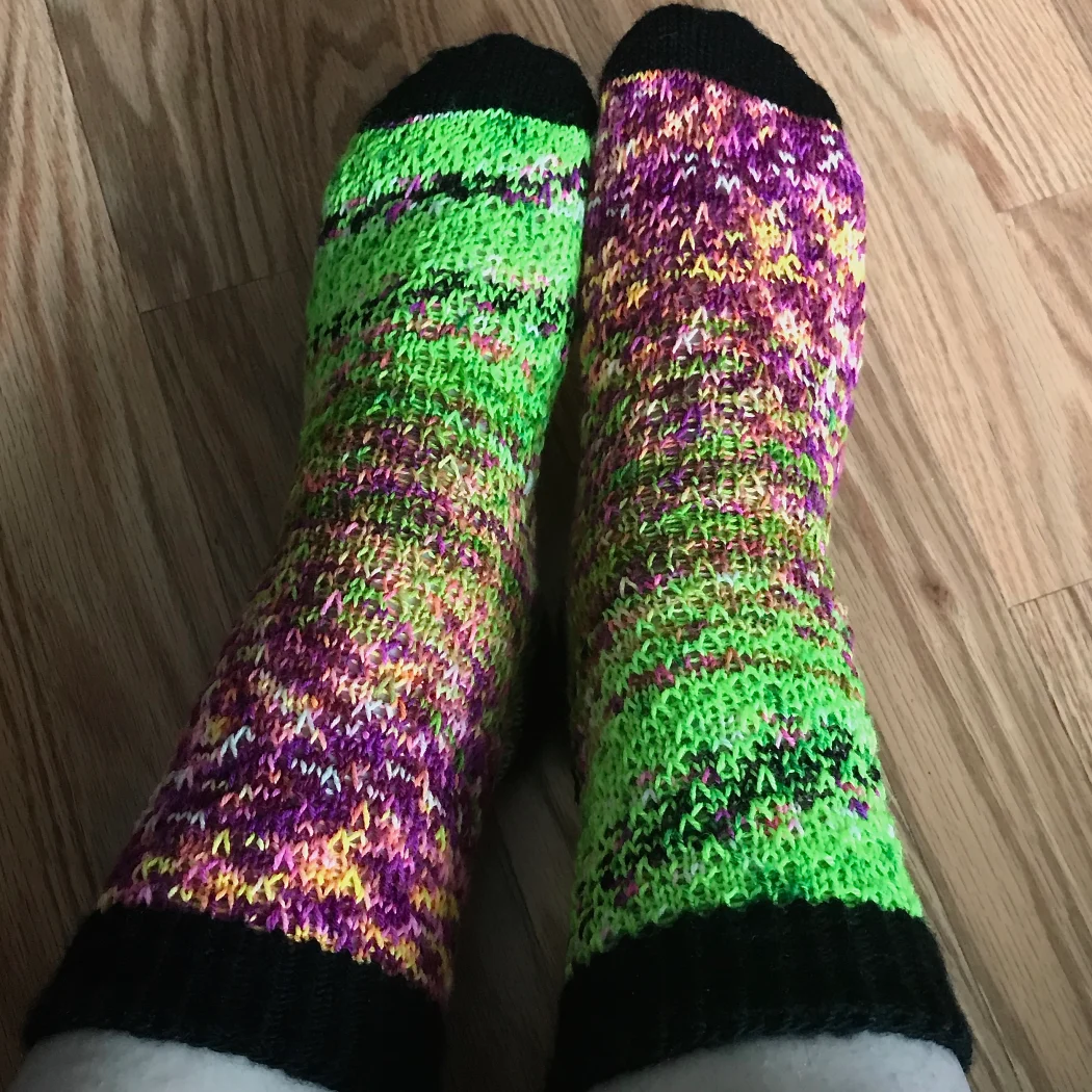 Top-down view of feet wearing green and purple socks with a fade effect with black heels, toes, and cuffs.