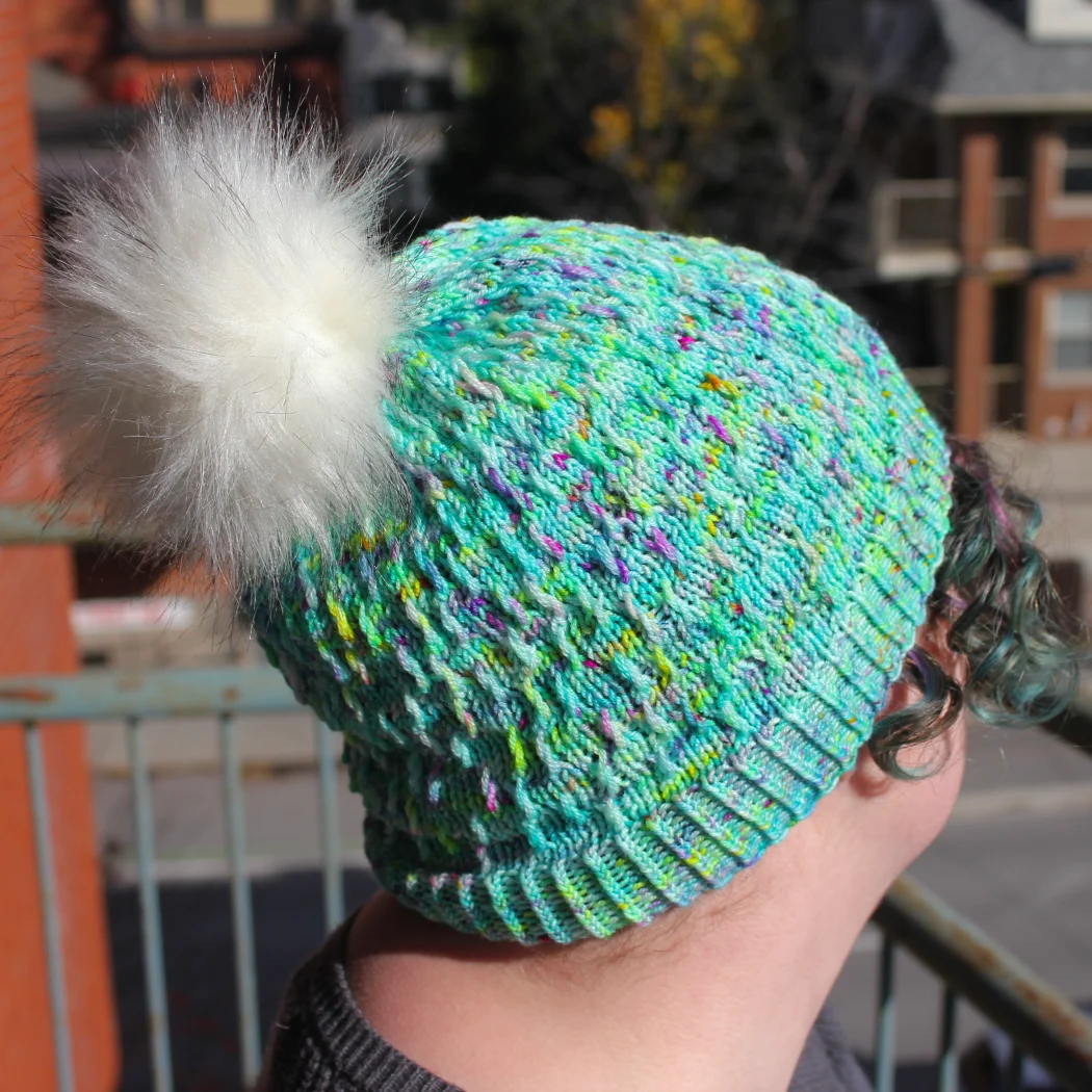 Side view of woman wearing bright green and rainbow hat with white pompom and twisted surface texture.