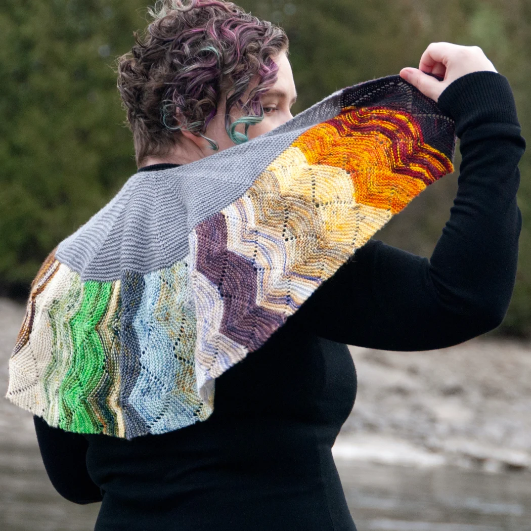 Woman wearing and holding a small, colourful crescent-shaped knitted shawl with undulating textured lines.