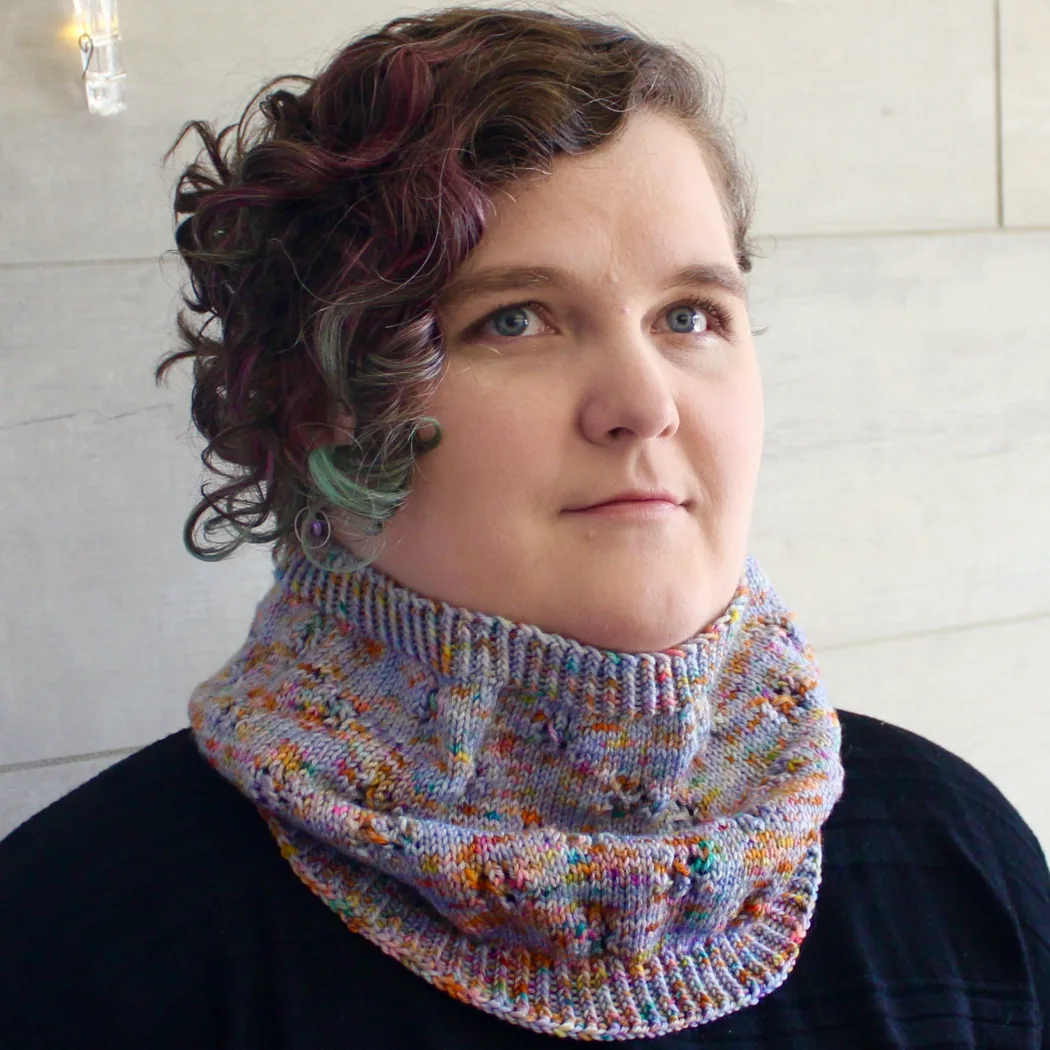 Woman wearing cowl that is light purple and rainbow-speckled knitted fabric with lace detail.