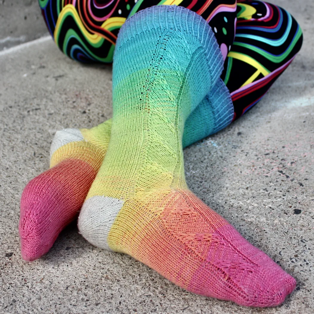 Crossed feet wearing rainbow socks with diagonal slipped-stitch panel and sparkly white heels.