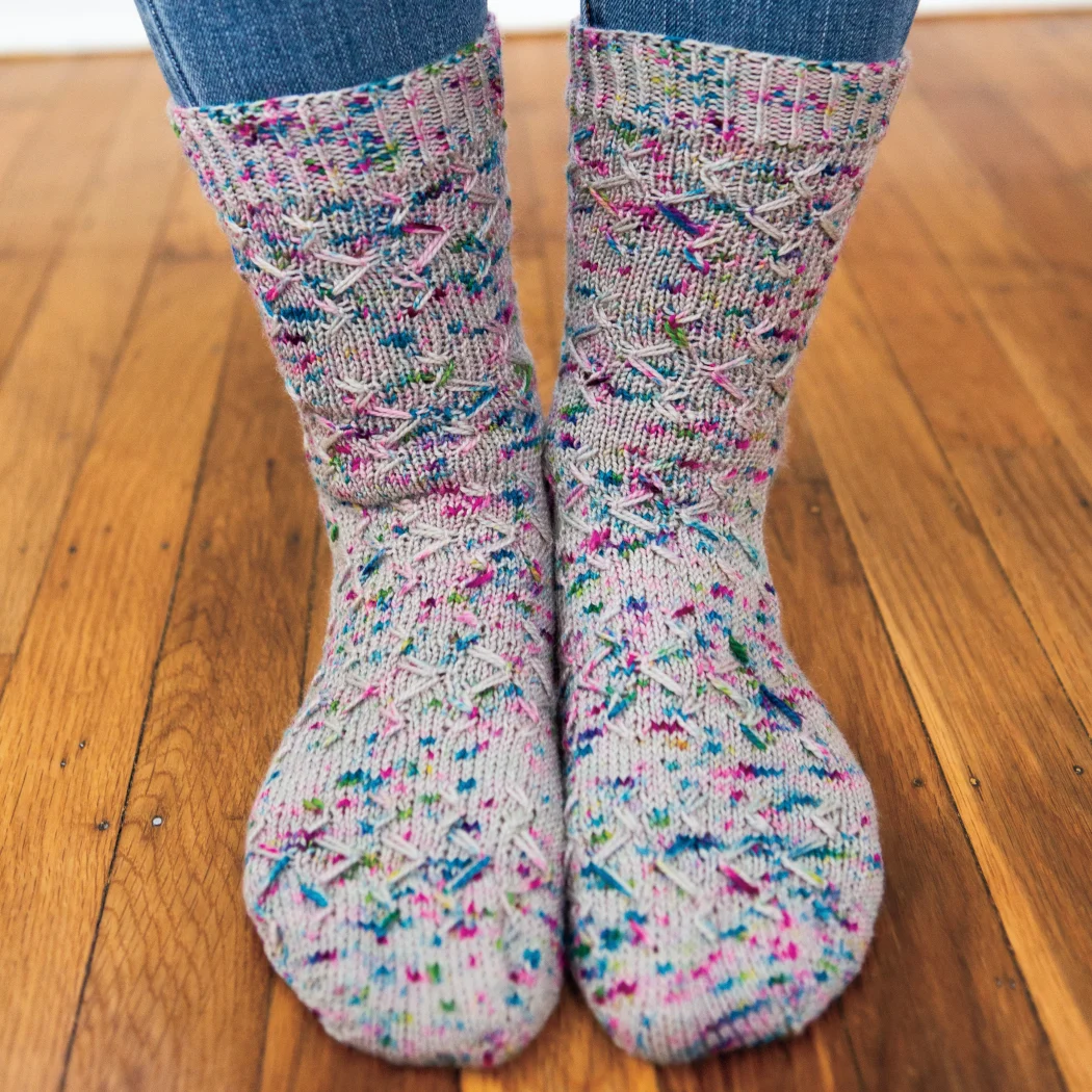 Front view of feet wearing grey knitted socks with blue and purple speckles and an angular slipped-stitch surface texture.